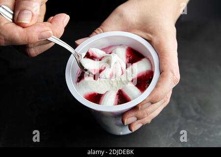 Yogurt ice cream in a white cup on black background, frozen sweet fruit dessert. Spoon with a cherry ice cream in female hand, closeup Stock Photo
