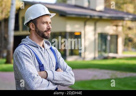 Close up of young male engineer in hard hat looking away, posing with arms crossed outdoors while working on cottage construction. Building, people, eco-friendly construction concept