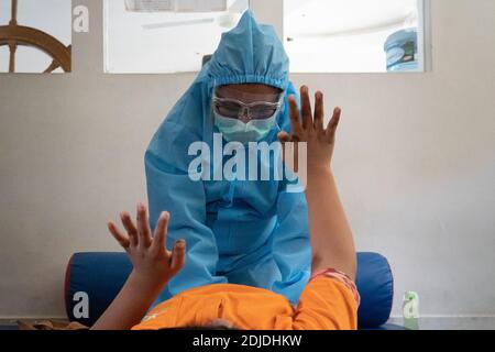 BALI/INDONESIA-OCT 23 2020: a child with a physical disability is doing physical therapy. They wear masks and hamzat suit to avoid the spread of the C Stock Photo