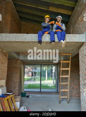 Two happy young builders in blue overalls and hard hats resting, sitting on the concrete floor, drinking coffee while working on house construction. Building house, partnership concept Stock Photo