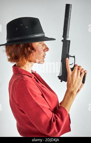 female with weapon against white background, isolated. detective redhead woman in black hat looking away, applying the weapon's barrel up Stock Photo