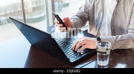 Remote work space. Freelancer work space near window. Man using laptop, holding smart phone for business work or study in smart watch drink water Stock Photo