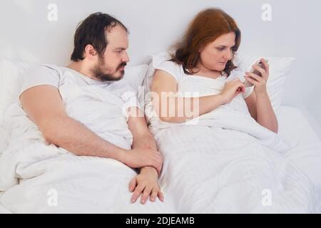 Husband looks wife phone while lying in white bed. A man peeks into a woman phone. Husband and wife spend time at home under the coronavirus quarantin Stock Photo