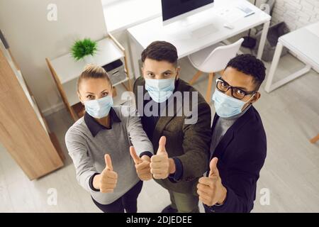 Young business people in medical face masks, looking at camera and giving thumbs-up Stock Photo
