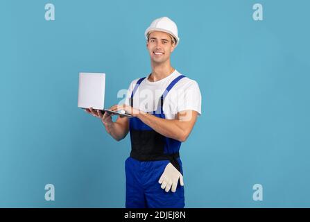 Man in coveralls and hard hat using laptop computer on blue background Stock Photo