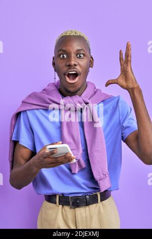 black man is in shock by news, while chatting with friend on smartphone, he is surprised, show emotions, gesturing with hands. isolated over purple ba Stock Photo