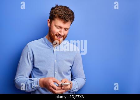 smiling bearded man is chatting with someone on smartphone, looks at screen of mobile phone, isolated over blue background Stock Photo