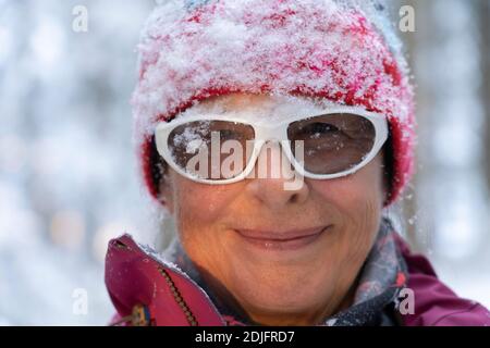 close up portrait of a pretty ageless senior woman in winter forest, standing under falling snow. Carefree woman enjoying snow and winter time. She pl Stock Photo