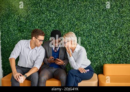 cheeful colleagues sit together in the aisle having leisure time, looking at screen of mobile phone smiling Stock Photo