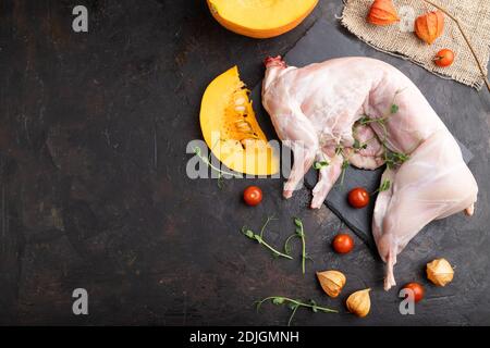 Whole raw rabbit with pumpkin, tomatoes, pea sprouts on a black concrete background and linen textile. Top view, flat lay, copy space. Stock Photo