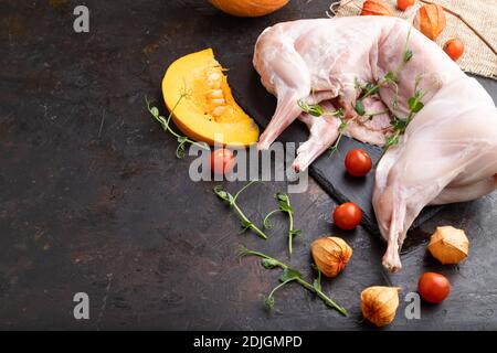 Whole raw rabbit with pumpkin, tomatoes, pea sprouts on a black concrete background and linen textile. Side view, copy space. Stock Photo