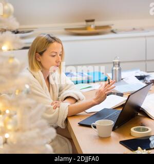 Young female entrepreneur wearing cosy warm bathrobe working remotly from home in winter Christmas time during 2020 corona virus pandemic. Work from Stock Photo