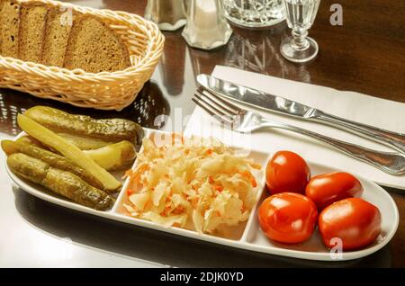 White dish with canned cucumbers, cabbage and tomatoes with black bread on the brown wooden table. Serving in restaurant. Stock Photo