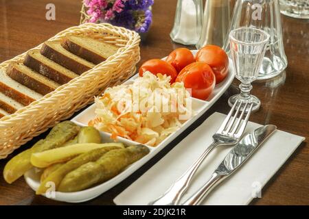 White large dish with canned cucumbers, cabbage and tomatoes, with black bread on the table of restaurant. Stock Photo