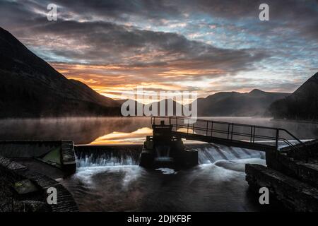 A beautiful winter sunrise at the weir at Crummock Water, with mist and a golden glow on clouds reflected in the lake. Lake District, Cumbria, England Stock Photo