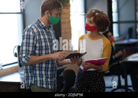 Two diverse creative colleagues wearing face masks talking in office Stock Photo