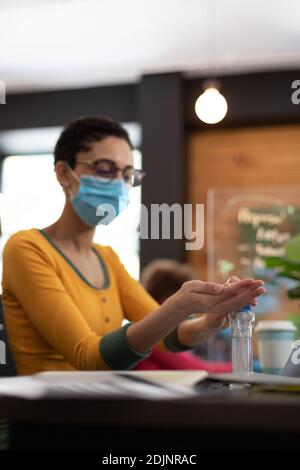 Mixed race woman wearing face mask sanitizing hands at desk in office Stock Photo