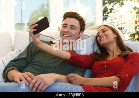 Caucasian couple taking selfie with smartphone sitting on couch smiling Stock Photo