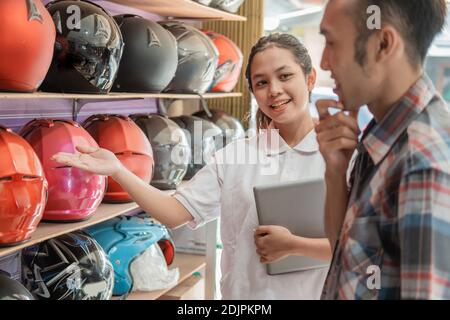 A woman's shop assistant holding a tablet in a hand gesture offers a helmet to the man in the helmet shop Stock Photo