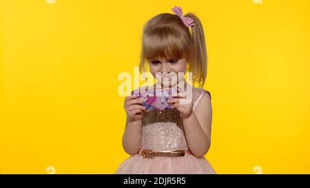 Technology for children. Little girl using smartphone. Portrait of child texting on smartphone. Kid enthusiastically playing games on mobile phone isolated on yellow background in studio. Stock Photo