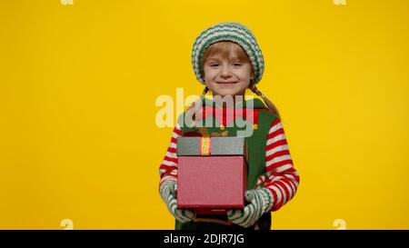Little teen kid girl in Christmas elf Santa helper costume isolated on yellow background. Child giving present gift box to camera, smiling sweetly. People New Year holidays celebration Stock Photo