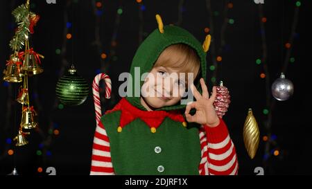 Blonde teen kid girl in Christmas elf Santa Claus helper costume showing okay sign isolated on black background with garland. Child smiles to camera with ok gesture. New Year holidays celebration Stock Photo