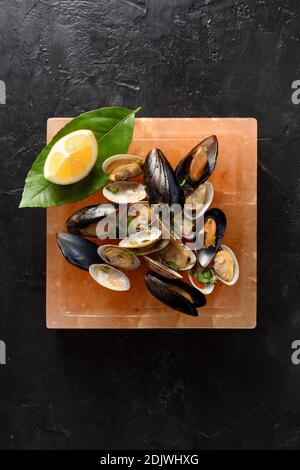 Salt block cooking. Vongole seafood mussels and clams in shells roasted on Himalayan pink salt block served with lemon on black background top view co Stock Photo