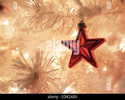 A shiny red star shaped ornament hangs in a white Christmas tree with bokeh and white twinkle lights during the holiday season. Stock Photo