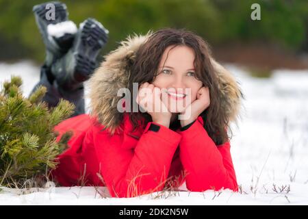 Nice brunette woman with long curly hair lies on winter snow in coniferous forest, smiling and looking to side Stock Photo