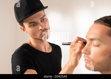 Concentrated young man in black cap doing theatrical makeup to man for photoshoot Stock Photo