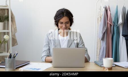 Happy young businesswoman self-employed tailor sitting at desk using laptop Stock Photo