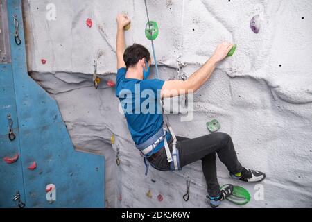 Active sporty man wearing protective face mask practicing rock climbing on artificial rock in a climbing wall. New normal in extreme sports. Stock Photo