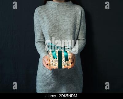 Woman in gray pullover holding luminous gift box in hands on dark background. Glowing yellow light bulbs into transparent gift box with dark green ribbon. Christmas and New Year concept Stock Photo