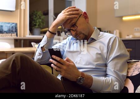 middle-aged man with puzzled look reading message or news item on his mobile phone, holding your head Stock Photo