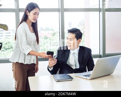 Asian businessman, manager in suit working with laptop computer looking at smartphone from young woman secretary on in office, working together for su Stock Photo