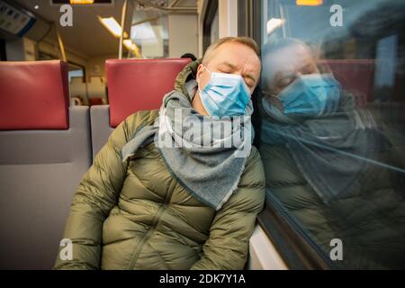 Traveling in public transport during the pandemic. Tired middle aged man wearing medical protective mask on his face sleeping in commuter train. Stock Photo