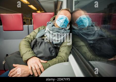 Traveling in public transport during the pandemic. Tired middle aged man wearing medical protective mask on his face sleeping in commuter train. Stock Photo