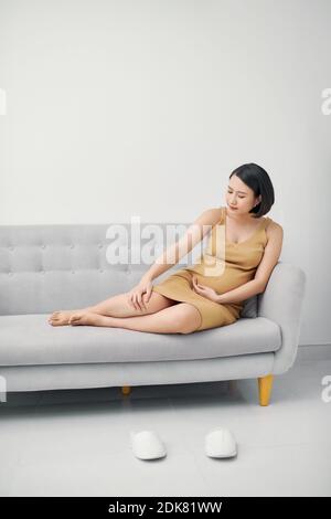 Young asian pregnant woman sitting on sofa having foot pain and leg cramps on her last trimester. Stock Photo
