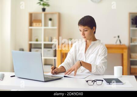 Young woman looking at laptop and making notes during online lesson Stock Photo