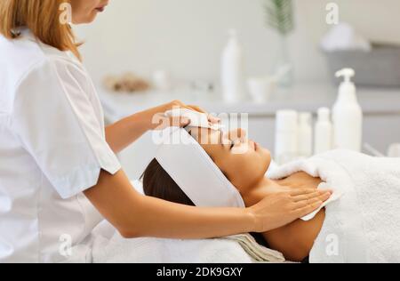 Cosmetologist wiping serene womans shoulders and forehead after facial massage in spa salon Stock Photo