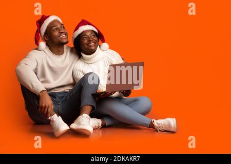 Dreamy african couple in santa hats sitting with laptop on orange background Stock Photo