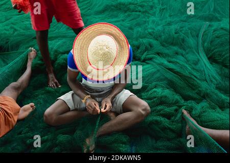 Unindentified fisherman, wearing bright hat, repairing large green fishing net with hands and feet in Kannur, Kerala, India. Stock Photo