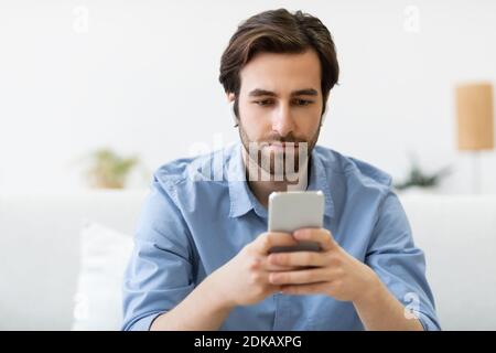 Serious Man Using Smartphone Wearing Earbuds Sitting On Sofa Indoors Stock Photo