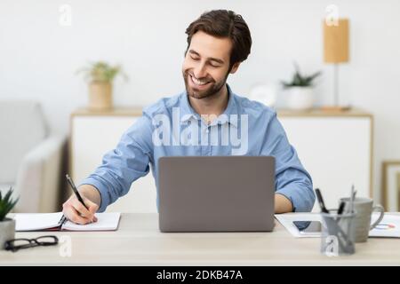 Cheerful Businessman Working At Laptop Taking Notes Sitting At Workplace Stock Photo