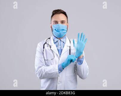 Confident young male doctor in facial mask putting on gloves over grey studio background Stock Photo