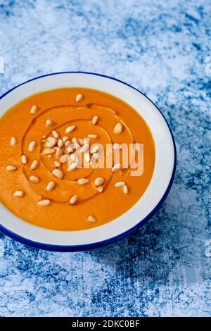 closeup of a white enamel bowl with pumpkin soup topped with some pine nuts and dressed with olive oil, on a blue granite surface Stock Photo