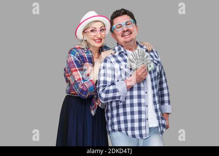 Happy wealthy family, adult man and woman in casual checkered shirt standing pickaback together, holding fan of dollar, toothy smile, looking at camer Stock Photo