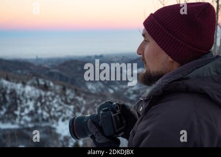 The guy with the camera, early in the morning in the winter in the mountains, looking out for the plot. The man is dressed in warm clothes of dark col Stock Photo