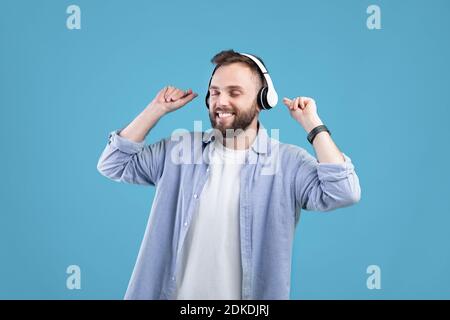 Happy young guy in headphones dancing to his favorite song, listening to music on blue studio background Stock Photo