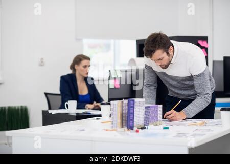 Young architect working on his construction model in the bright office with his colleague in the background. Stock Photo
